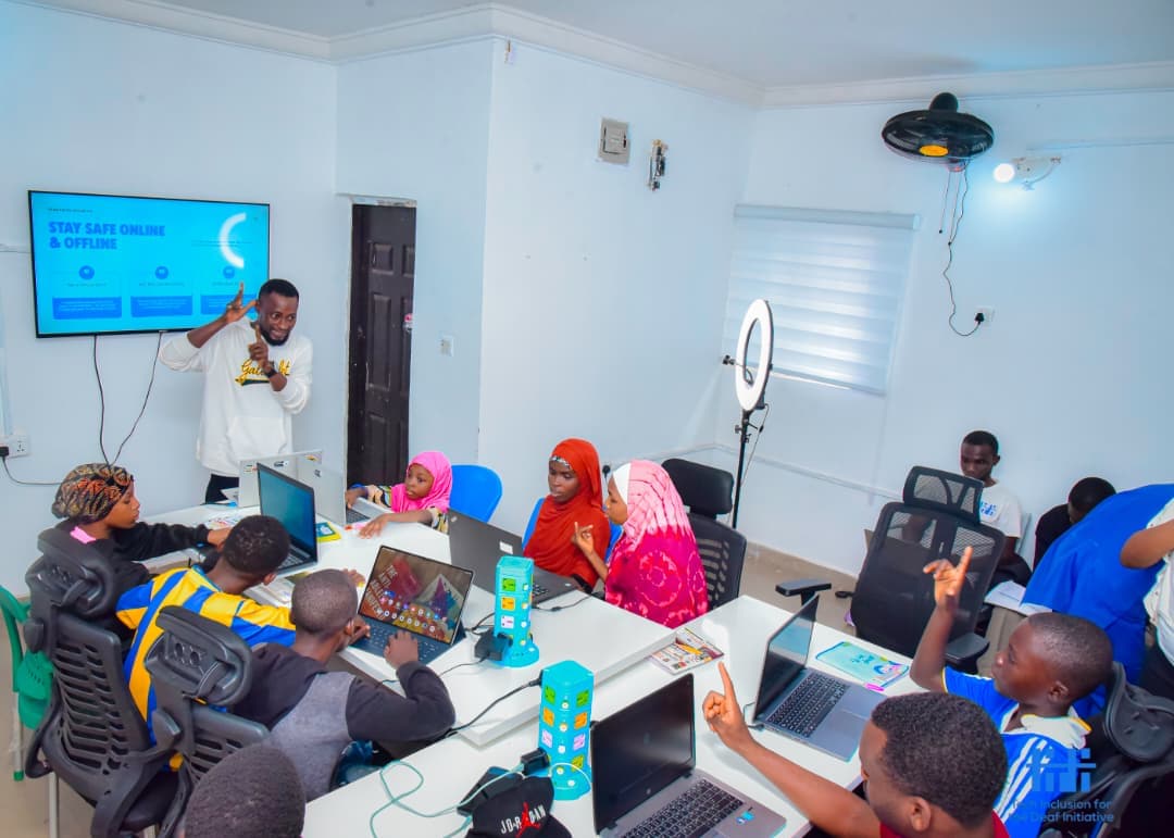 A diverse group of Nigerian children and teens, including girls wearing colorful hijabs, sit at white tables with laptops in a bright tech classroom. A male instructor in a white sweatshirt stands at the front teaching with hand gestures. A large TV screen on the wall displays "STAY SAFE ONLINE & OFFLINE". The room has white walls, office chairs, a ring light, and ceiling fan. Some students are raising hands and engaging during the TIDI Tech Bootcamp.